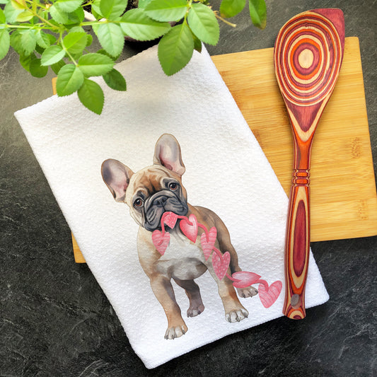 a french bulldog with a pink ribbon on a tea towel next to a wooden spoon